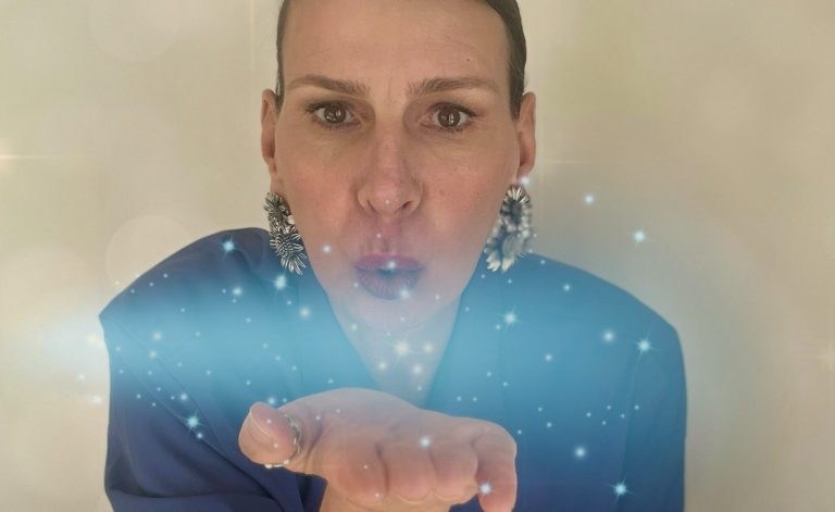 Ellen mit großen Ohrringen bläst funkelnde Lichtpartikel in die Kamera.