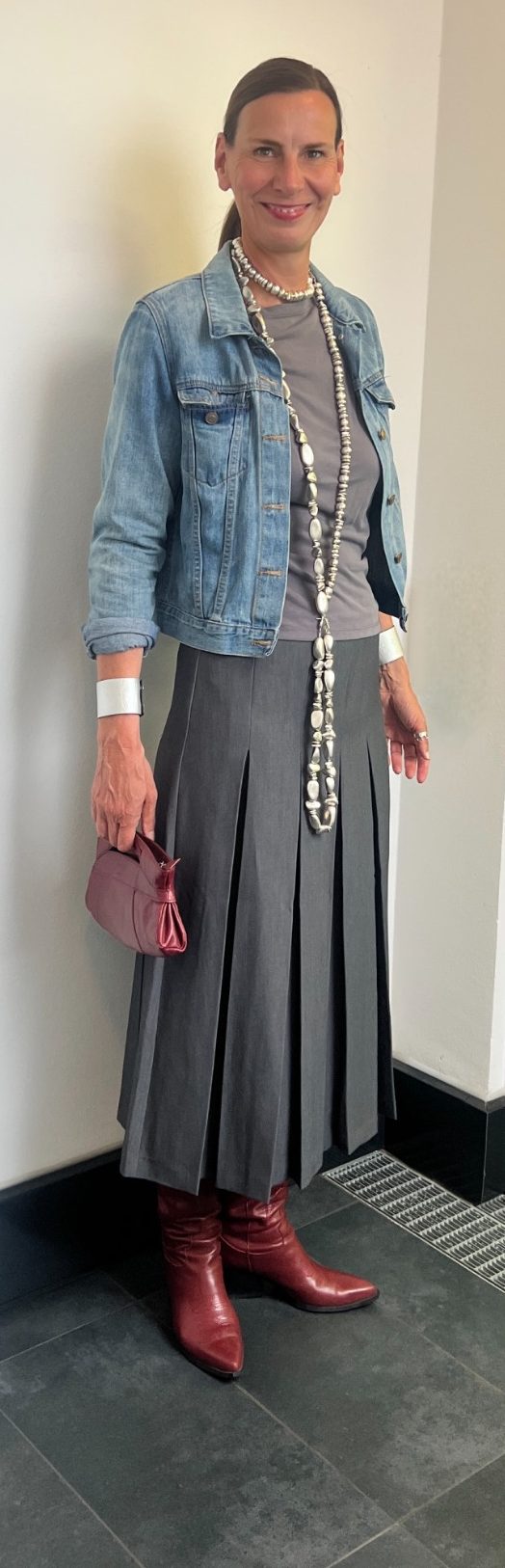 Gut aussehen. Gut fühlen. Ellen Schmitt in Jeansjacke, grauem Kleid und roten Stiefeln, mit einer Handtasche.