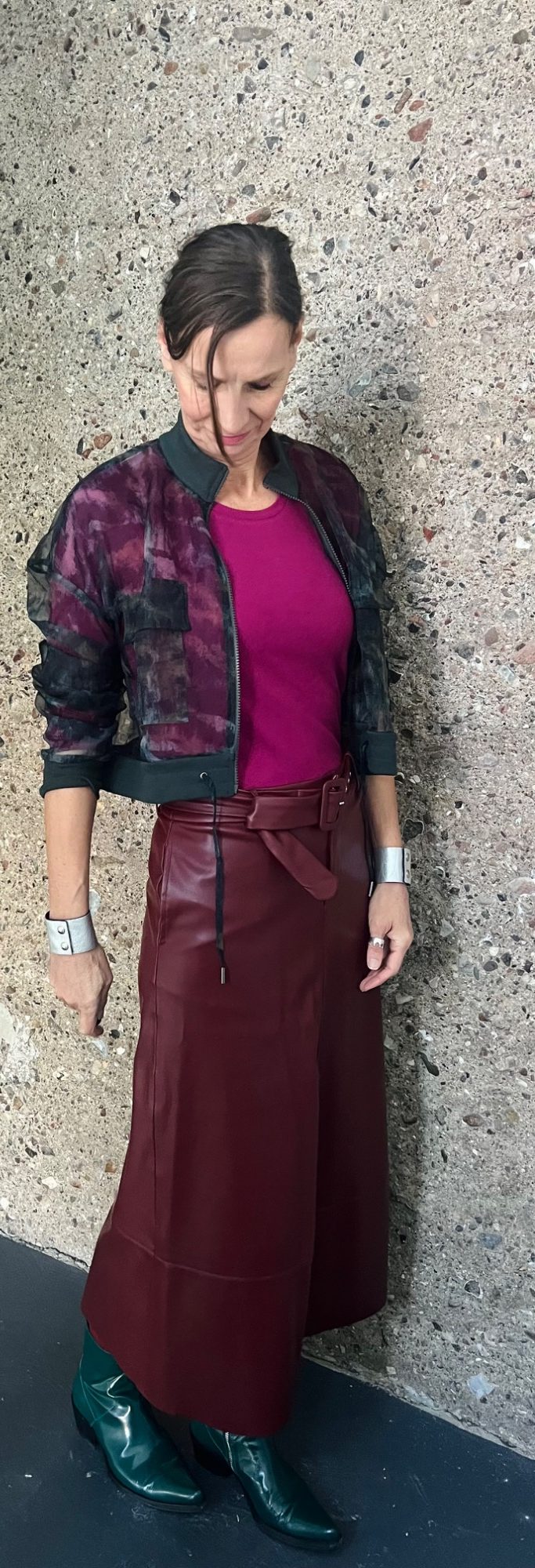 Gut anziehen. Gut aussehen. Gut fühlen. Ellen Schmitt in rot-violettem Oberteil und brauem Rock, steht vor einer grauen Wand.