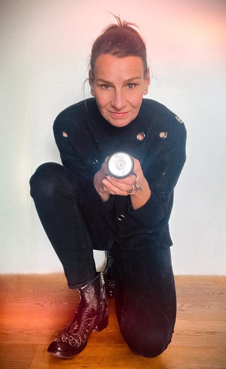 Ich erhöhe Deine Strahlkraft Ellen Schmitt in Schwarz hält eine Taschenlampe und posiert kauernd auf einem Holzfußboden.
