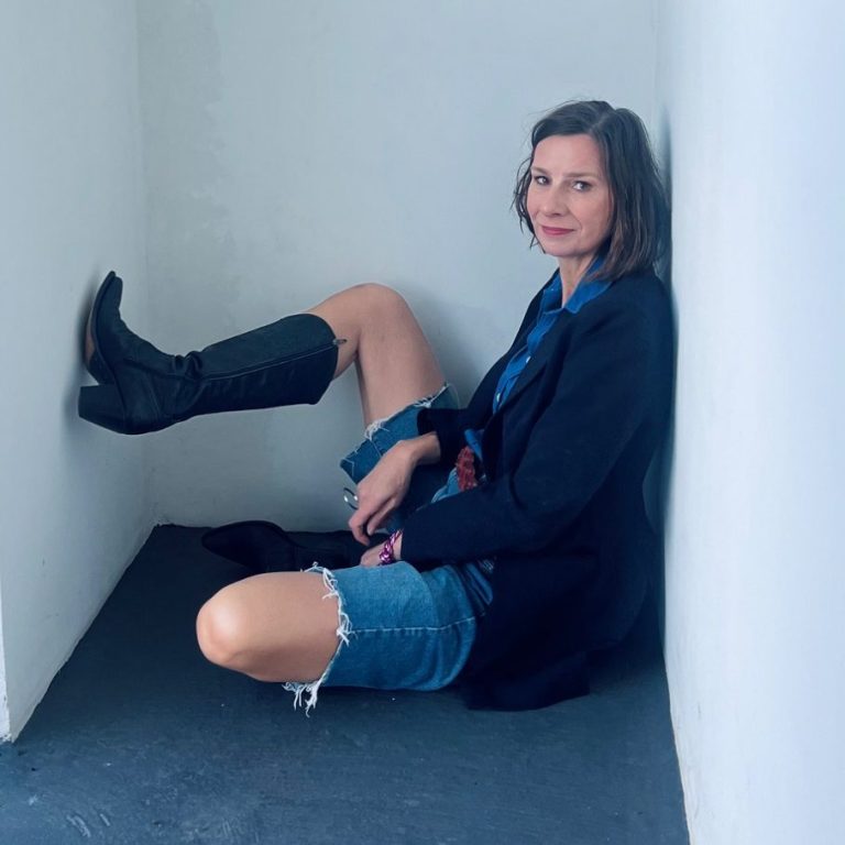 Strahlen mit Stil Frau sitzt in einer Ecke, trägt Lederstiefel, Blazer und eine kurze Jeans, entspannte Pose.