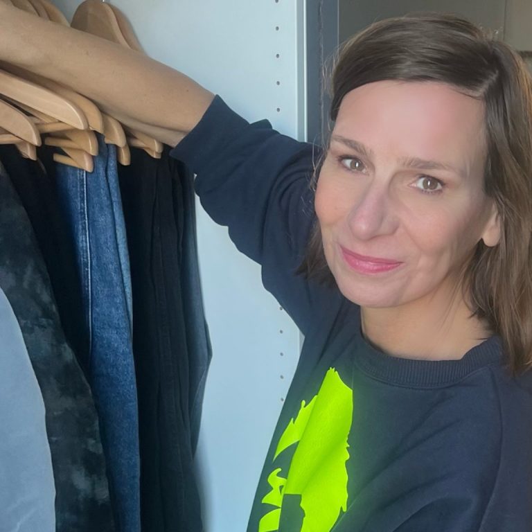 Strahlkraft-Outfit Ellen Schmitt in blauem Sweatshirt zeigt auf Kleiderständer mit verschiedenen Kleidungsstücken.