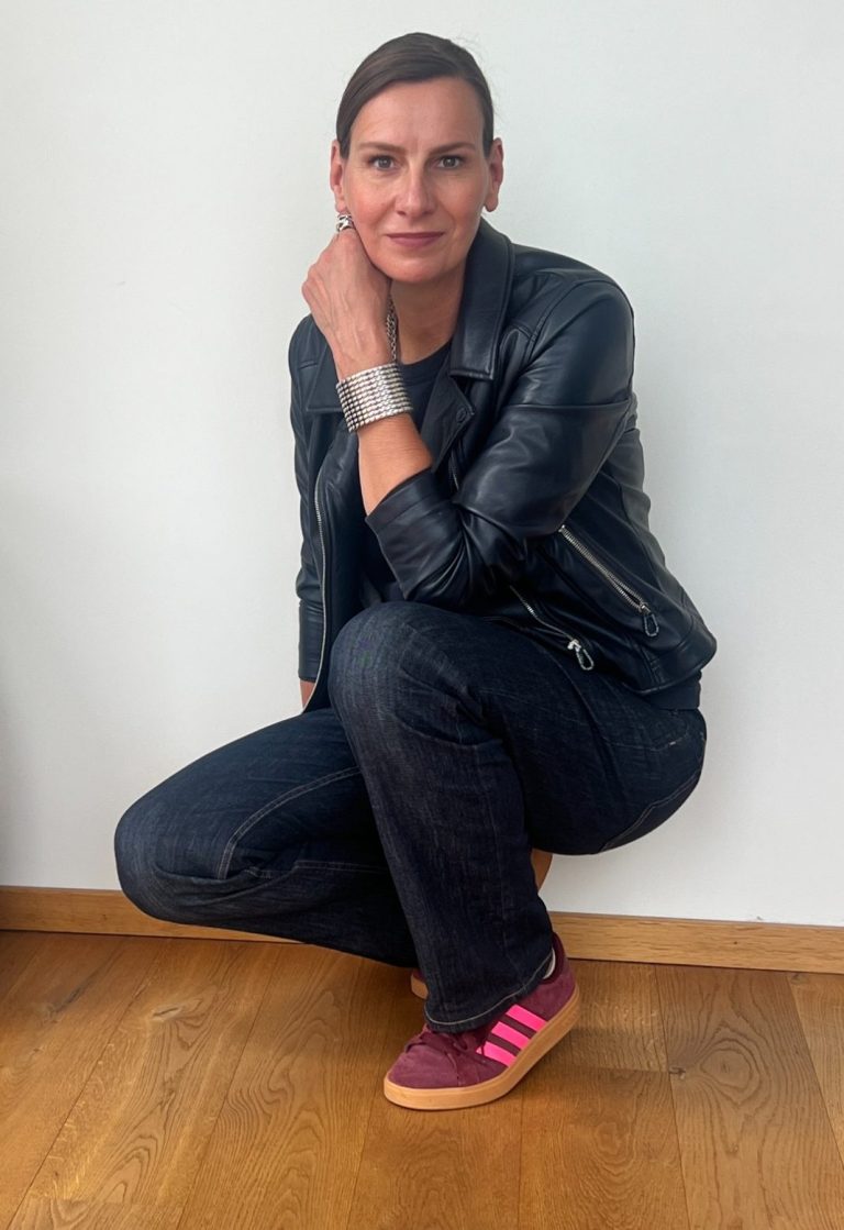 Strahlkraft-Trainerin Ellen Schmitt in Lederjacke und Jeans kniet mit einem Handgelenkarmband auf dem Boden.